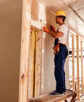 Handsome young man in safety helmet and work overalls painting wall with paint roller